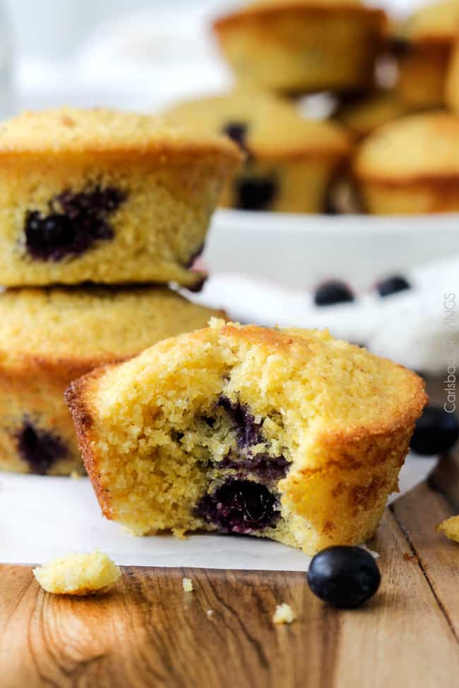 close up of a blueberry cornbread muffin with bite missing. Two muffins are stacked in background. 