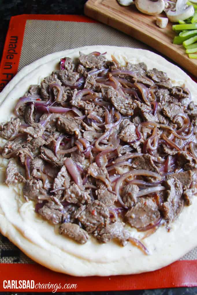 showing how to make  Philly Cheesesteak Pizza by layering Alfredo sauce with cheese, steak and onions