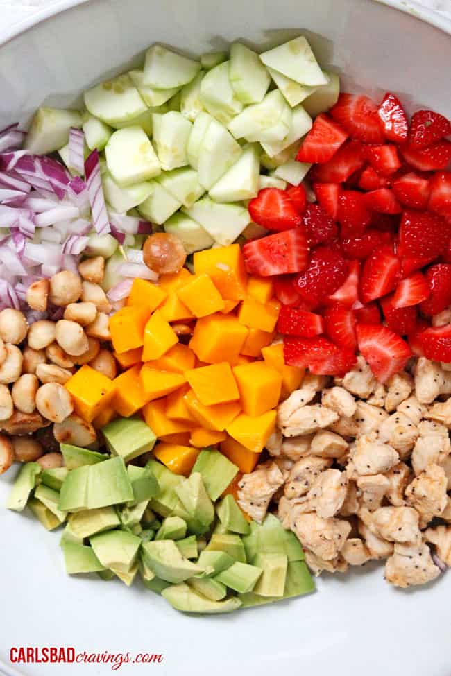 top view of showing how to make chicken fruit salad by adding chicken, avocados, mangos, cucumbers, red onions and macadamia nuts to a bowl