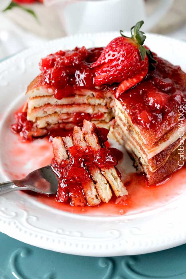 a stack of strawberry cheesecake pancakes on a white plate. A chunk has been cut out and is speared on a fork. 