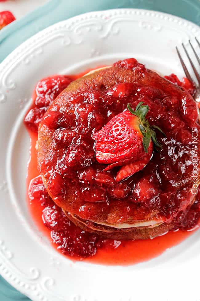 overhead view of stack of strawberry pancakes on a white plate that's resting on a blue cake stand. 