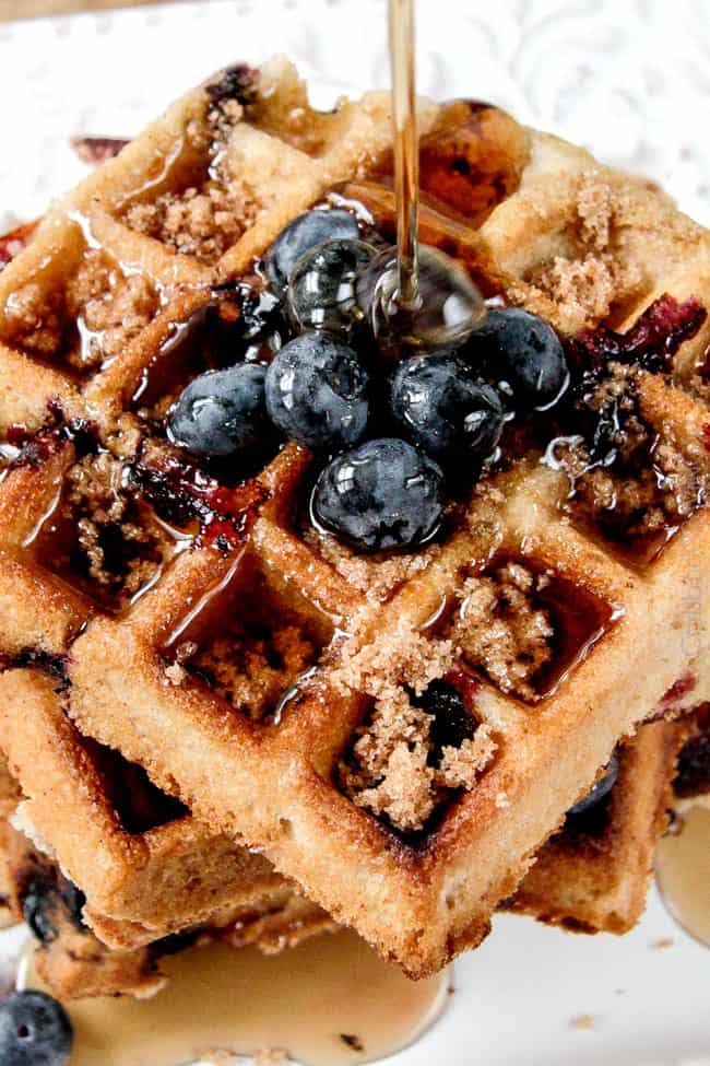 Top view of a stack of four Blueberry Waffles on a white plate with blue berries and syrup being poured on top. 