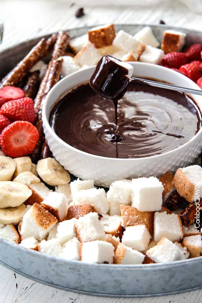 pulling up a marshmallow out of dark Chocolate Fondue recipe surrounded by a platter o dippers with bananas, strawberries, pretzels and pound cake