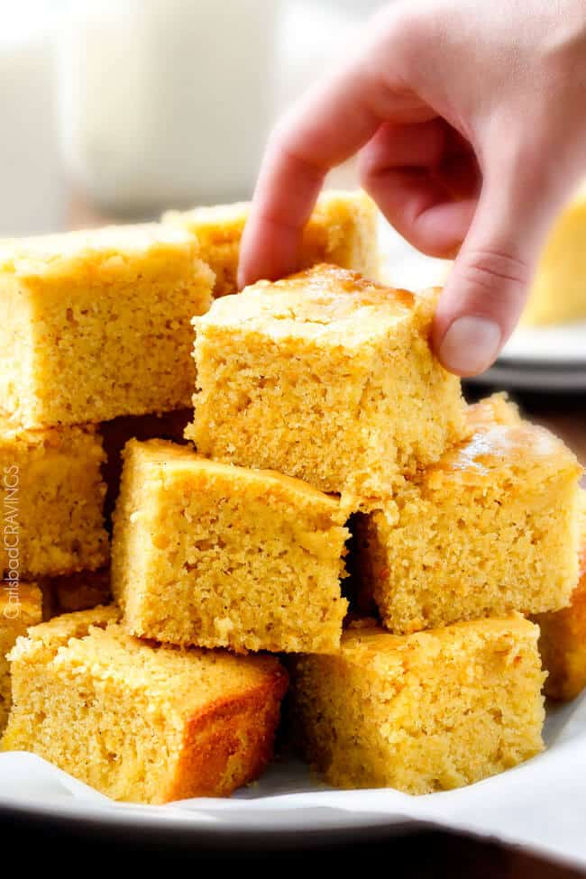 a hand picking up a slice of Sweet Cornbread stacked on a plate