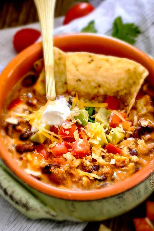 up close of a bowl of taco soup with a spoon in in i t topped with sour cream, tomatoes, cheese, avocados and tortilla chips