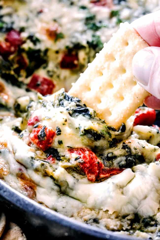 up close of a cracker scooping cheesy spinach dip