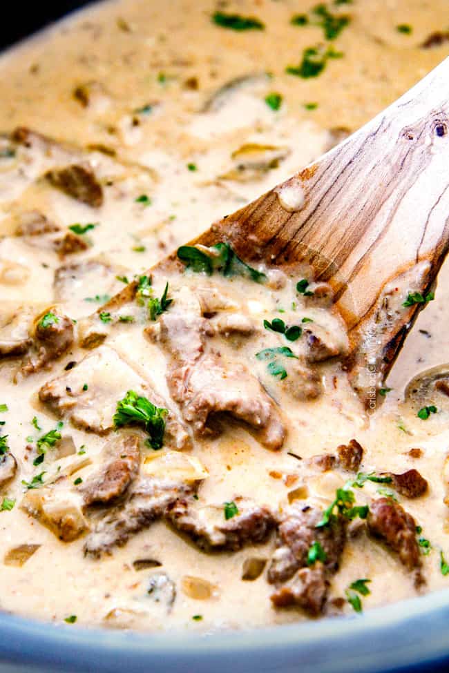 up close of crock pot Beef Stroganoff in the crock pot with a wooden spoon in rich gravy garnished with parsley