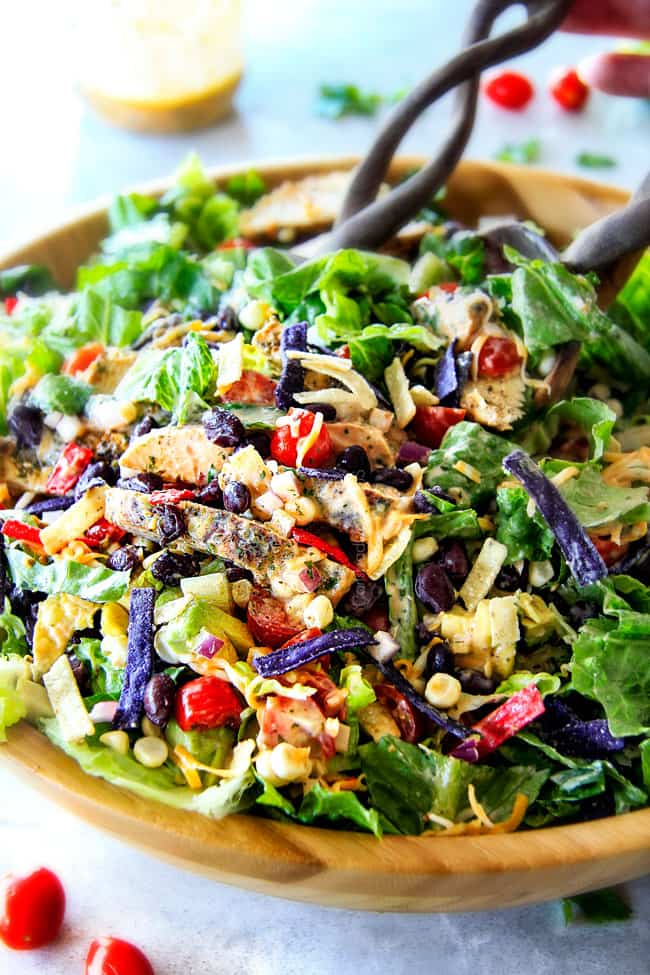 showing how to make chicken taco salad by tossing the chicken, black beans, corn, bell peppers, avocados, tomatoes, cheese, tortilla chips and dressing together in a wooden bowl