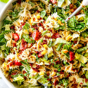 Top Shot of Best BLT Pasta Salad in a white bowl with white tongs lifting the salad up