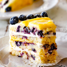slice of lemon blueberry cake with a piece taken out of it sitting on a white plate