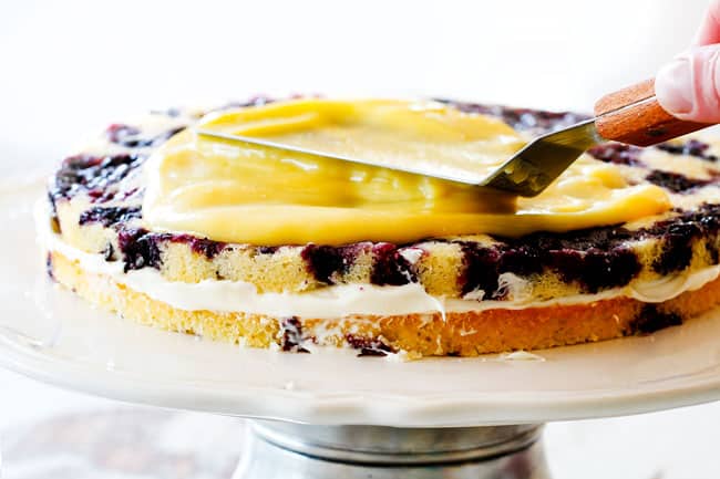 Frosting Lemon Blueberry Cake with Lemon Curd on a white cake pedestal