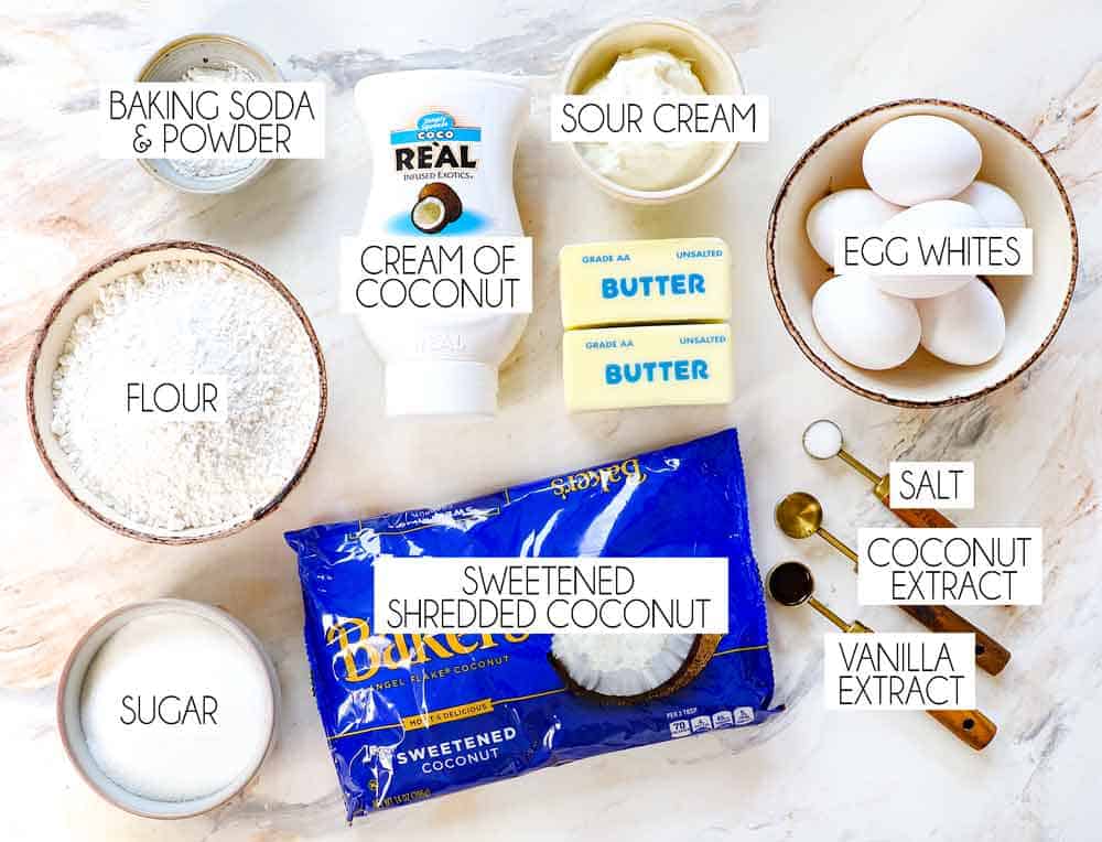 top view of coconut cake ingredients: flour, baking powder, baking soda, sugar, coconut, sour cream, butter, eegg whites, vanilla extract, coconut extract, salt and cream of coconut