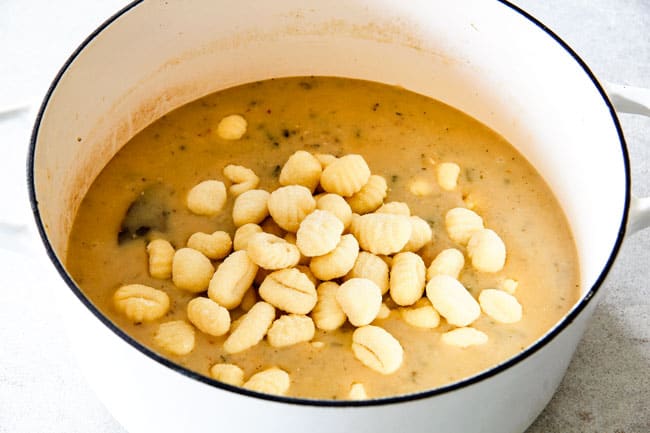 Showing how to make healthy Chicken Gnocchi Soup by adding potato gnocchi to soup in a white dutch oven