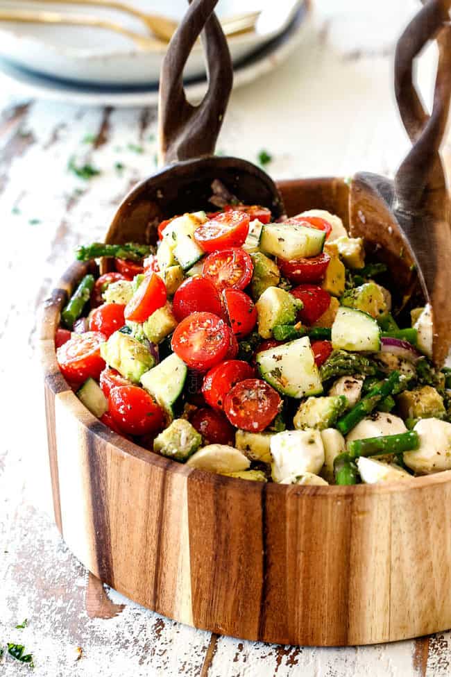 far away shot of Cucumber Tomato Salad recipe with cherry tomatoes, cucumbers, onion, asparagus and avocado in a wood bowl with wood tongs scooping salad