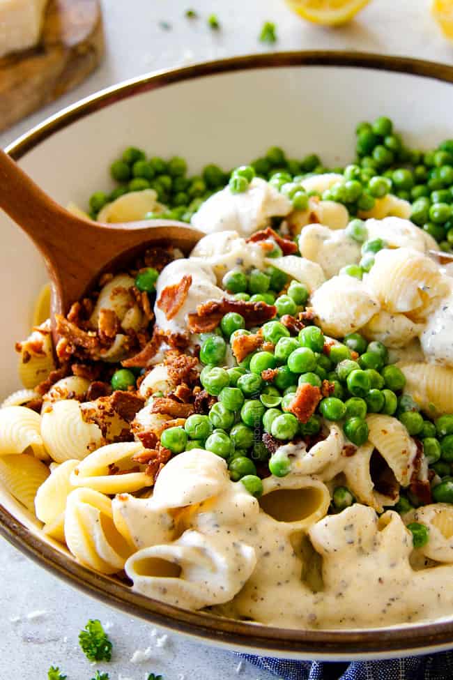 showing how to make Creamy Pasta Salad by adding salad ingredients of bacon and peas to a white bowl