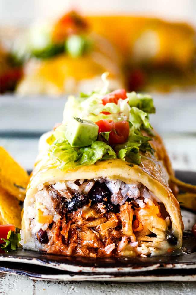 front view of wet burrito cut open with Mexican Chicken, rice, black beans on a black plate garnished with lettuce, tomatoes, sour cream