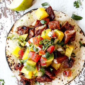 up close top view of al pastor pork on a corn tortilla with grilled pineapple, pico de gallo and avocado on a white board