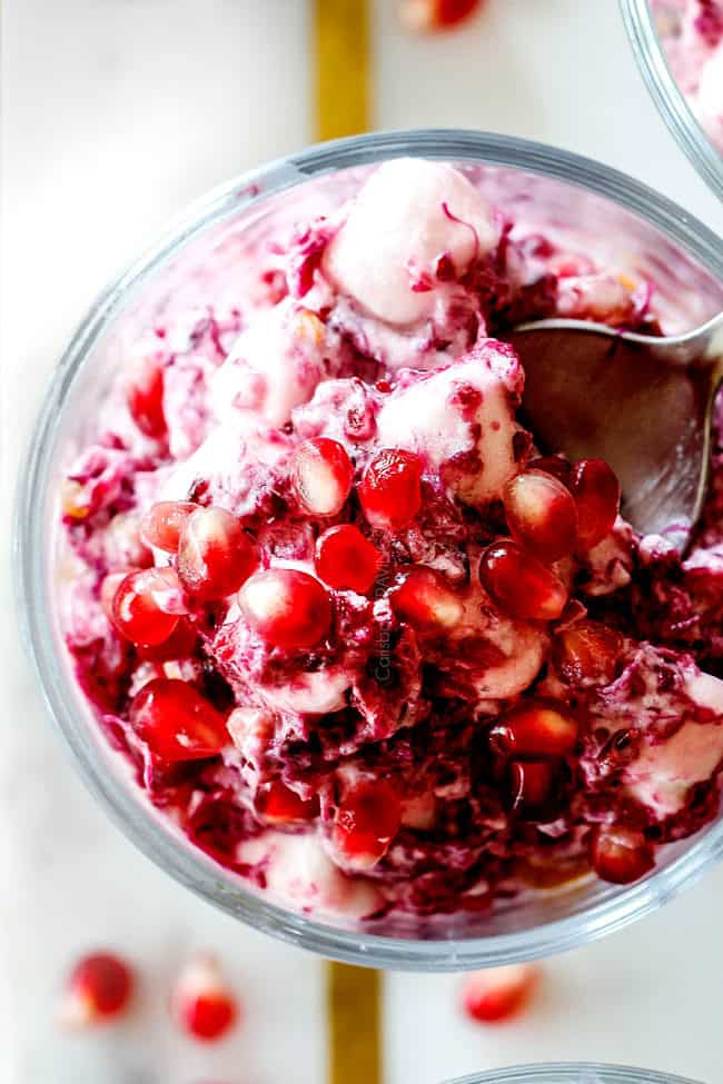 up close top view of a spoonful of creamy ambrosia salad with pineapple, mandarin oranges, mini marshmallows, cranberries and pomegranate seeds with creamy dressing with yogurt in small glass serving dishes
