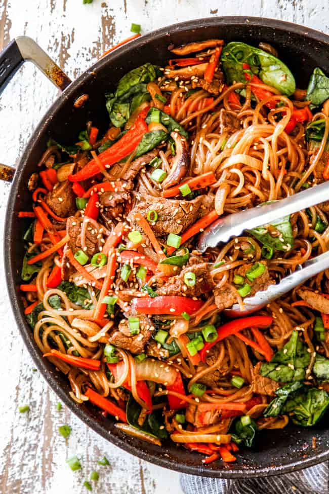 top view of spicy Korean noodles with tongs grabbing glass noodles, steak and vegetables