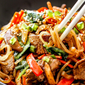 a bowl of Korean Spicy Noodles with beef, mushrooms, carrots, sesame seeds, green onions with white chopsticks