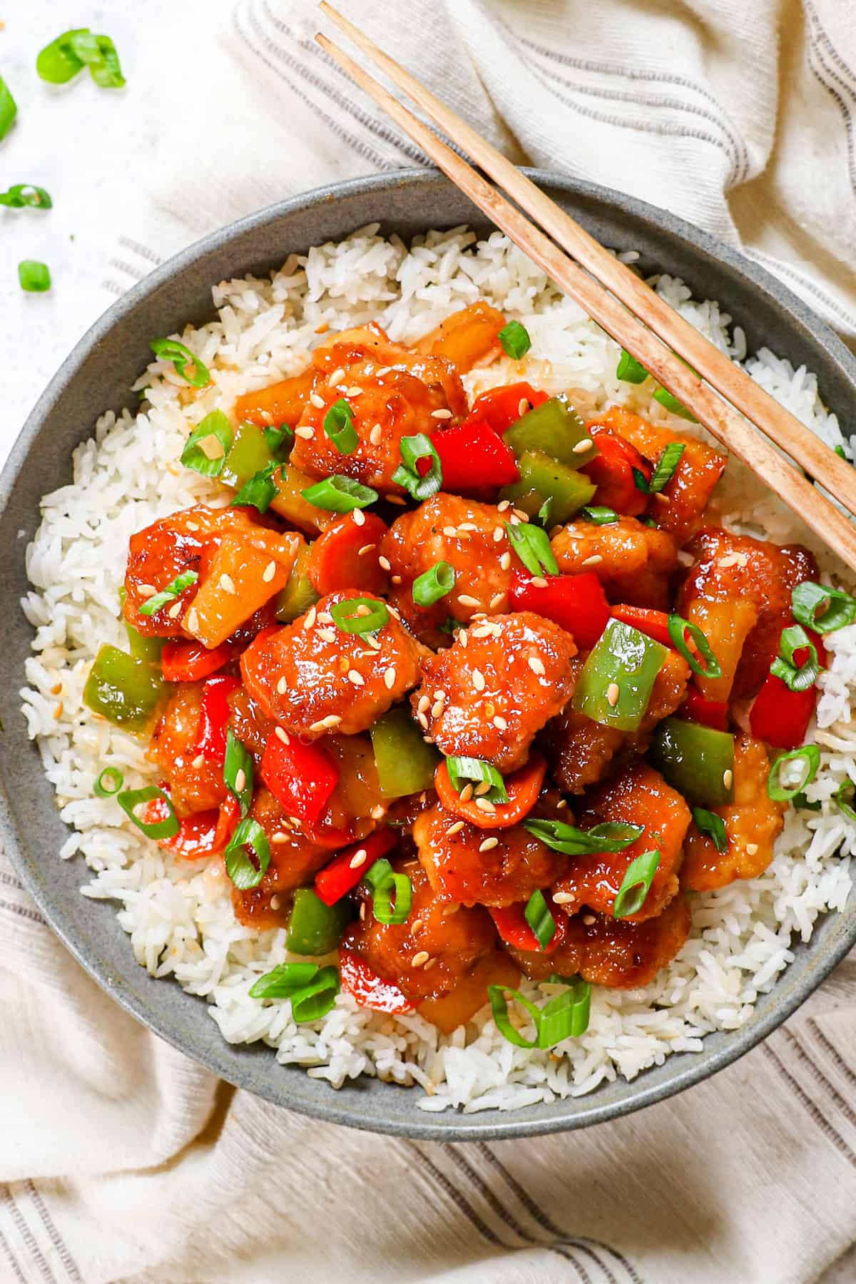 showing how to served baked sweet and sour chicken by adding to a bowl with rice, garnishing with sesame seeds, green onions and using chopsticks