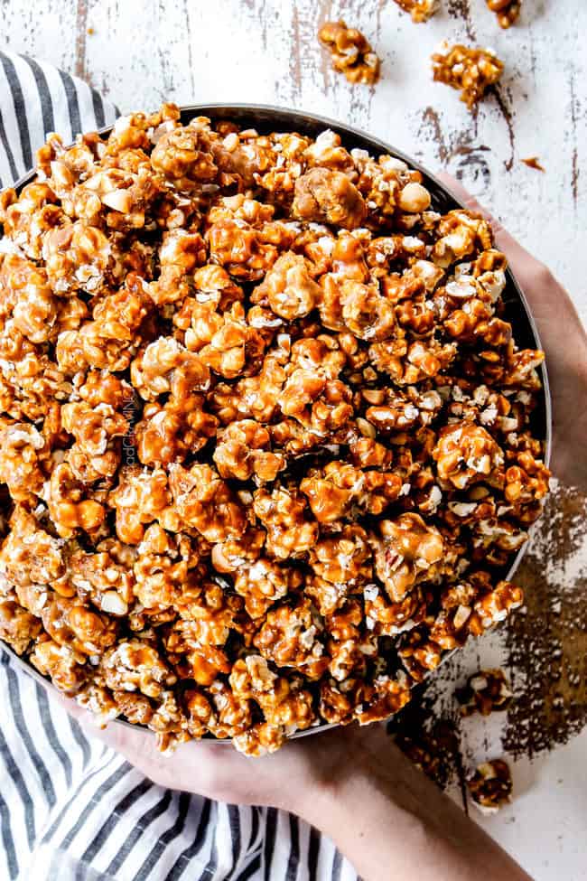 two hands holding a large bowl of easy caramel popcorn in a silver bowl