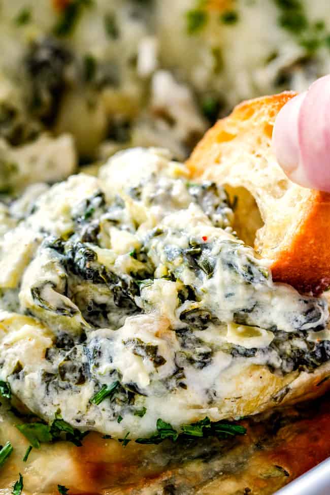 up close of a piece of bread dipping into spinach and artichoke dip