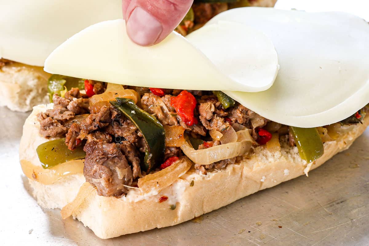 showing how to make Philly cheesesteak by adding slices of provolone over the steak mixture on the bun