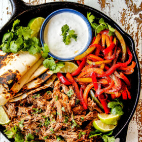 top view of pork fajitas in a skillet with salsa verde pork, bell peppers, onions, limes, cilantro and tortillas