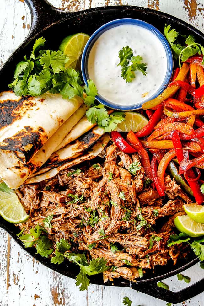 up close of top view of pork fajitas in a skillet with salsa verde pork, bell peppers, onions, limes, cilantro and tortillas