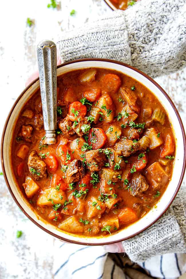 two hands holding a bowl of old fashioned beef stew showing what's in it:  beef, potatoes, carrots, celery, onions, garlic, gravy