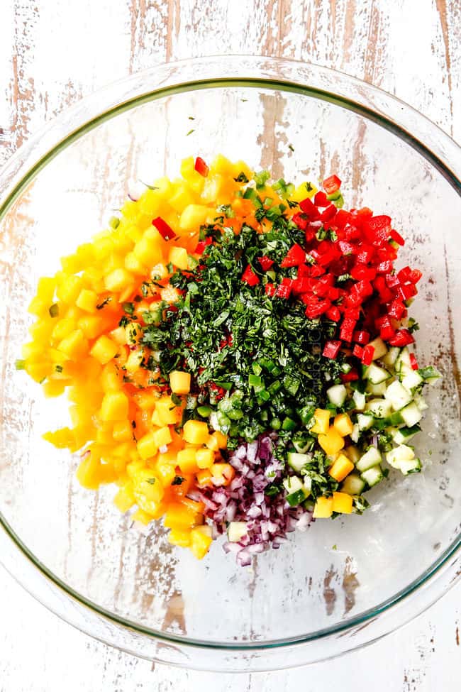 mostrando come fare la salsa di mango per il salmone con vista dall'alto di una ciotola di vetro con mango, jalapenos, cilantro, peperoni rossi, cipolle rosse