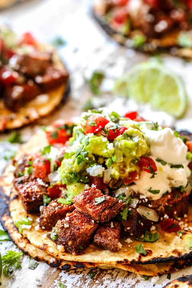 showing how to assemble marinated carne asada tacos by layering with carne asada, sour cream, guacamole, pico de gallo