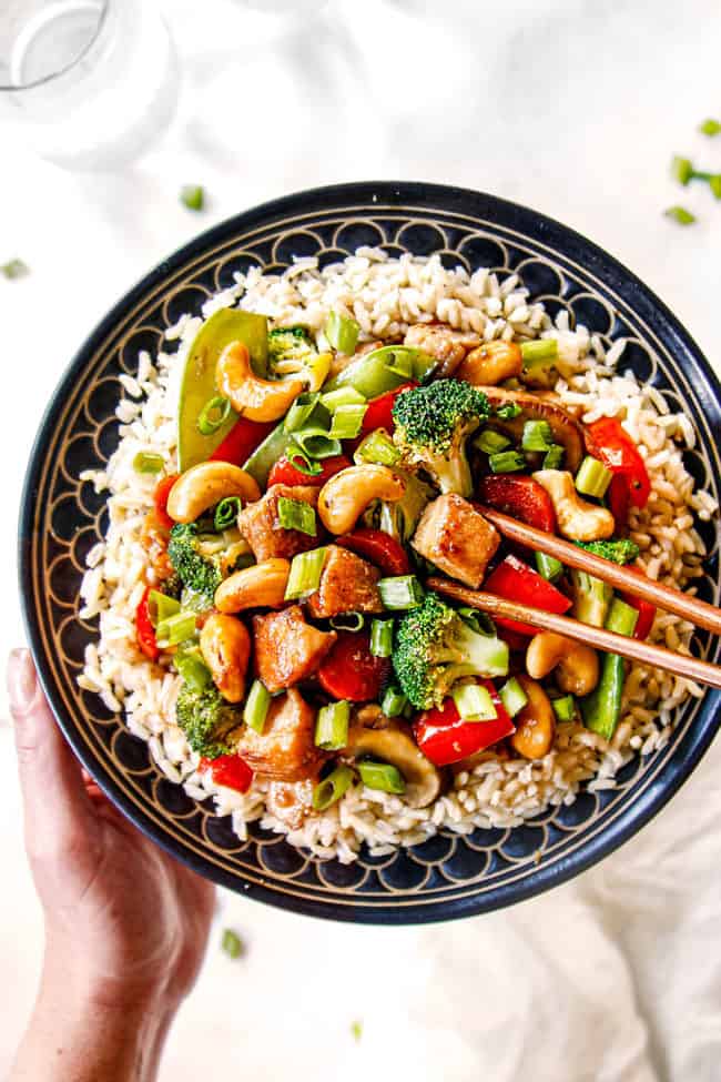 two hands holding a bowl of chicken and broccoli stir fry recipe over rice