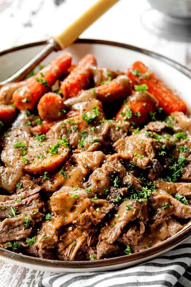 showing how to serve slow cooker pot roast by adding shredded chuck roast, carrots and potatoes to a white bowl and drizzling with gravy