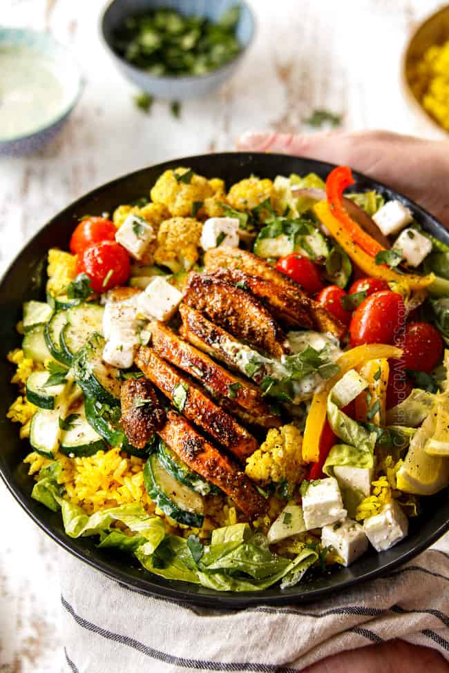 showing to make chicken shawarma into bowls by adding chicken, roasted cauliflower, bell peppers, tomatoes and zucchini to a black bowl with rice and lettuce