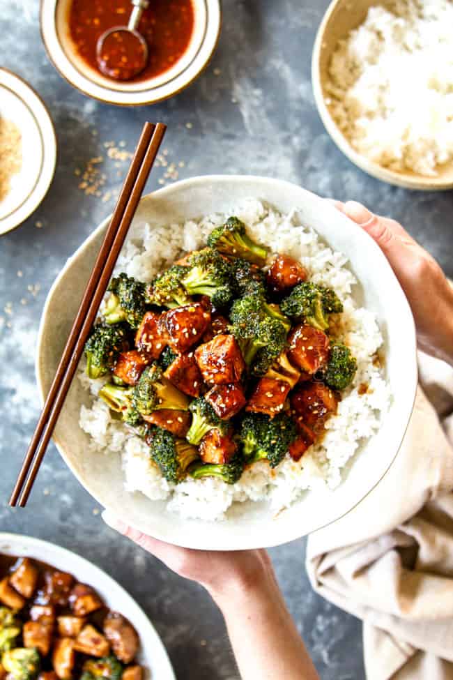 showing how to serve Chinese chicken and broccoli recipe with two hands holding a bowl of chicken and broccoli over rice with chopsticks
