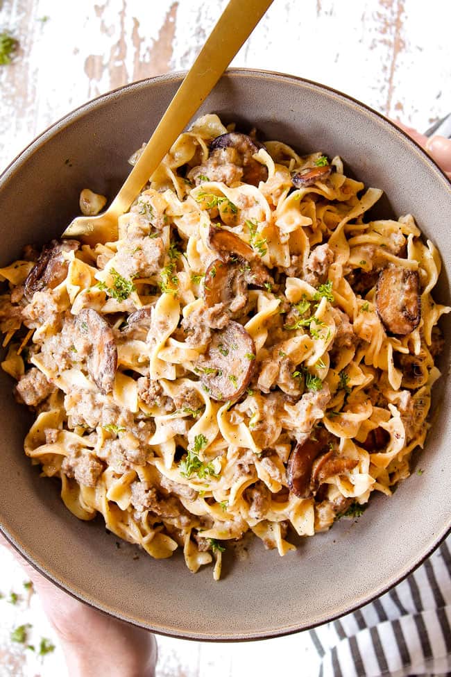 top view of two hands holding a bowl of beef stroganoff with ground beef over egg noodles showing what to serve stroganoff with