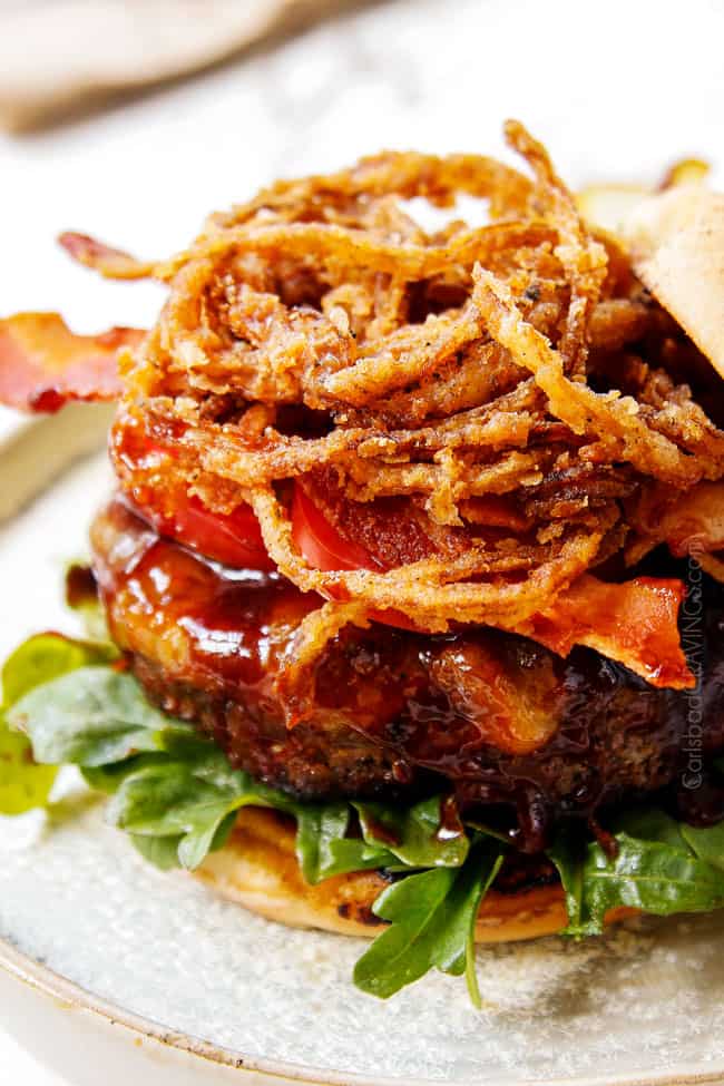 showing how to make BBQ burger recipe by adding a pile of bacon and fried onion strings on top of burger before adding the top bun