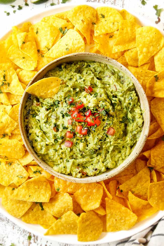 showing how to garnish guacamole recipe by topping with fresh tomatoes in  a tan bowl surrounded by tortilla chips