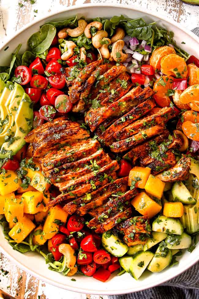 top view showing how to make mango salad by adding mangos, lettuce, chicken, cucumbers, red bell peppers and cashews to a salad bowl