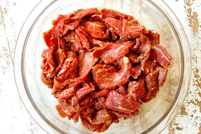 showing how to make Pepper Steak  by marinating slices of flank steak in a glass bowl with soy sauce, sesame oil, rice wine, Chili sauce and cornstarch 