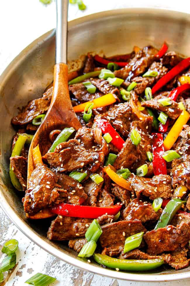 showing how to make Chinese Pepper Steak by combining steak,  bell  peppers and stir fry sauce in a stainless steel pan  with a wooden spoon