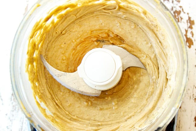 showing how to make hummus recipe by blending tahini, lemon juice, garlic cloves, olive oil, ground cumin and salt to a food processor until smooth