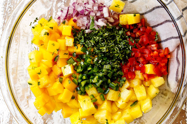 showing how to make steak taco recipe by making salsa by adding pineapple, mangos, jalapenos, red onion and cilantro to a glass bowl