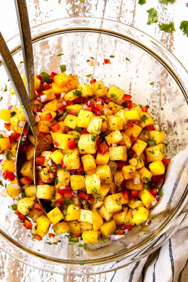 showing how to make top view of pineapple mango salsa by tossing pineapple, mangos, red onions, red bell peppers, cilantro and jalapeno together in a glass bowl