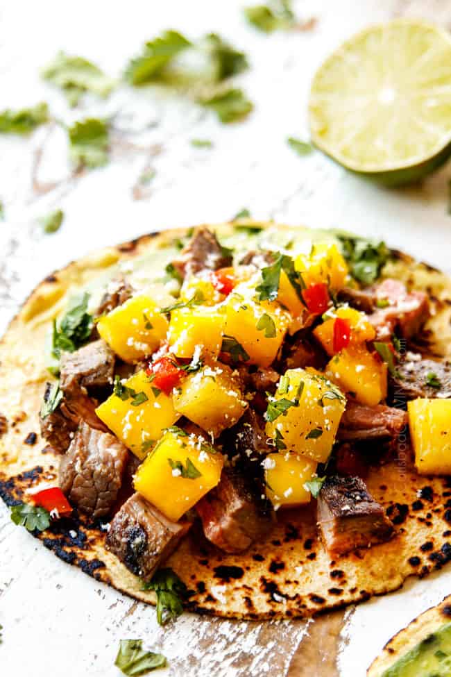 showing how to make flank steak tacos by layering corn tortillas with flank steak and pineapple mango salsa