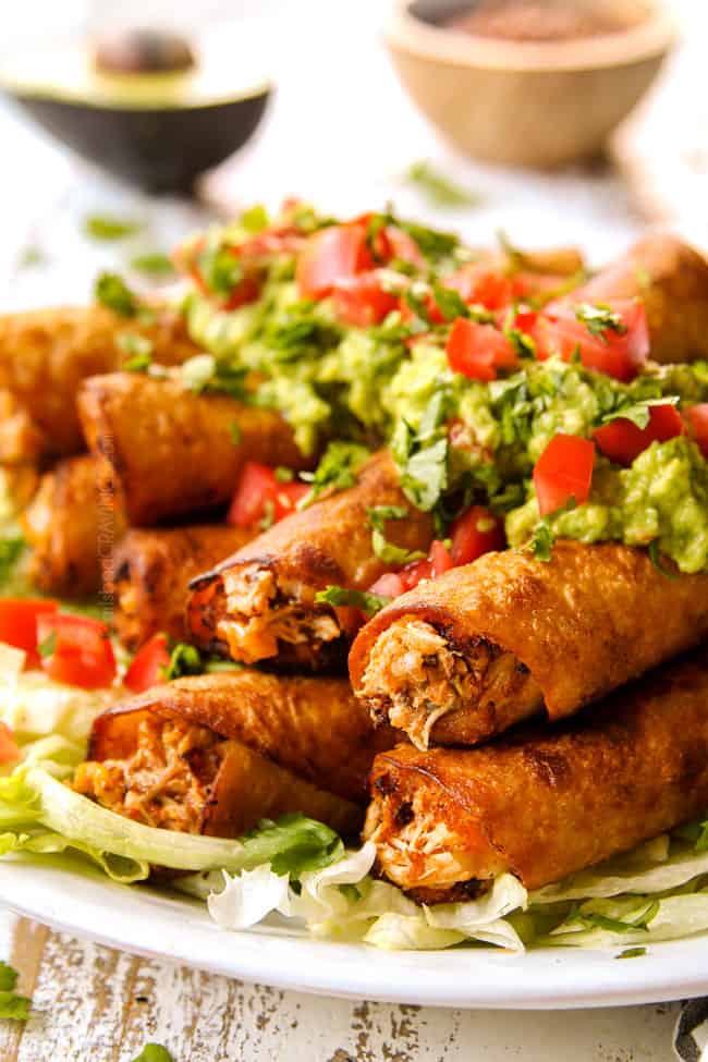 side view of crispy fried or baked flautas stacked on a white plate with shredded lettuce, guacamole and chopped tomatoes