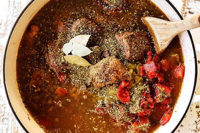 showing how to make  pozole by adding chicken broth, diced tomatoed, Mexican oregano, ground cumin and bay leaves to the pork in a white Dutch oven