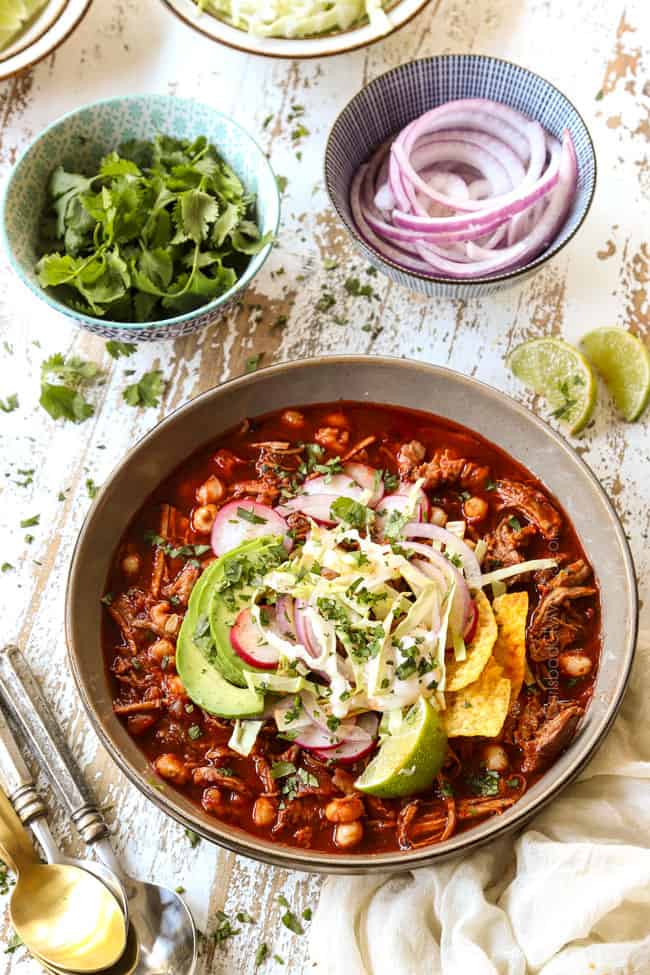 a bowl of pozole rojo surrounded by bowls of garnishes: cilantro, limes, red onions, radishes
